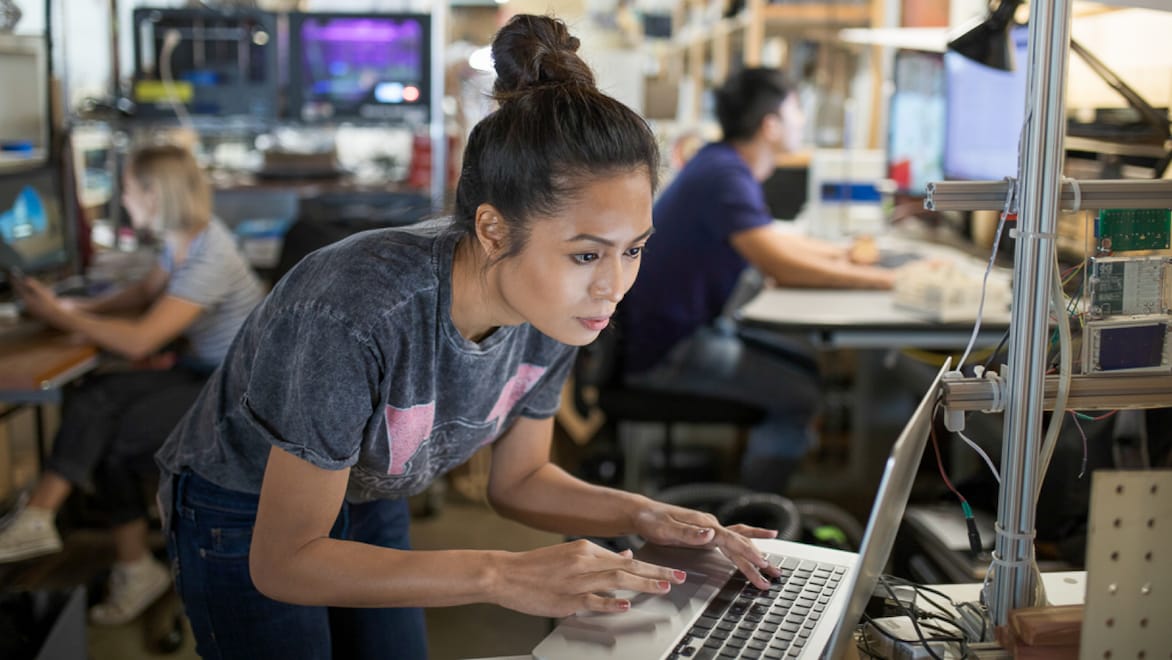 Woman working at a laptop in a computer lab. Another woman and man working at separate computers in the background.